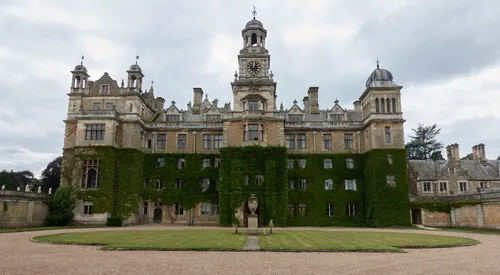 The image is an architectural photograph capturing the entrance of Thoresby Hall, a grand country house located in Nottinghamshire, England, United Kingdom. The photo was taken in the late afternoon during the early summer, as indicated by the bright yet slightly overcast sky and the lush green ivy climbing the building’s facade. Thoresby Hall itself serves as the focal point, showcasing its ornate Victorian architecture, central clock tower, and extensive stonework, all of which are hallmarks of this historic landmark. The well-maintained lawns and the gravel path at the forefront further enhance the stately appearance of this English country house, making it a prominent feature in the Nottinghamshire landscape.